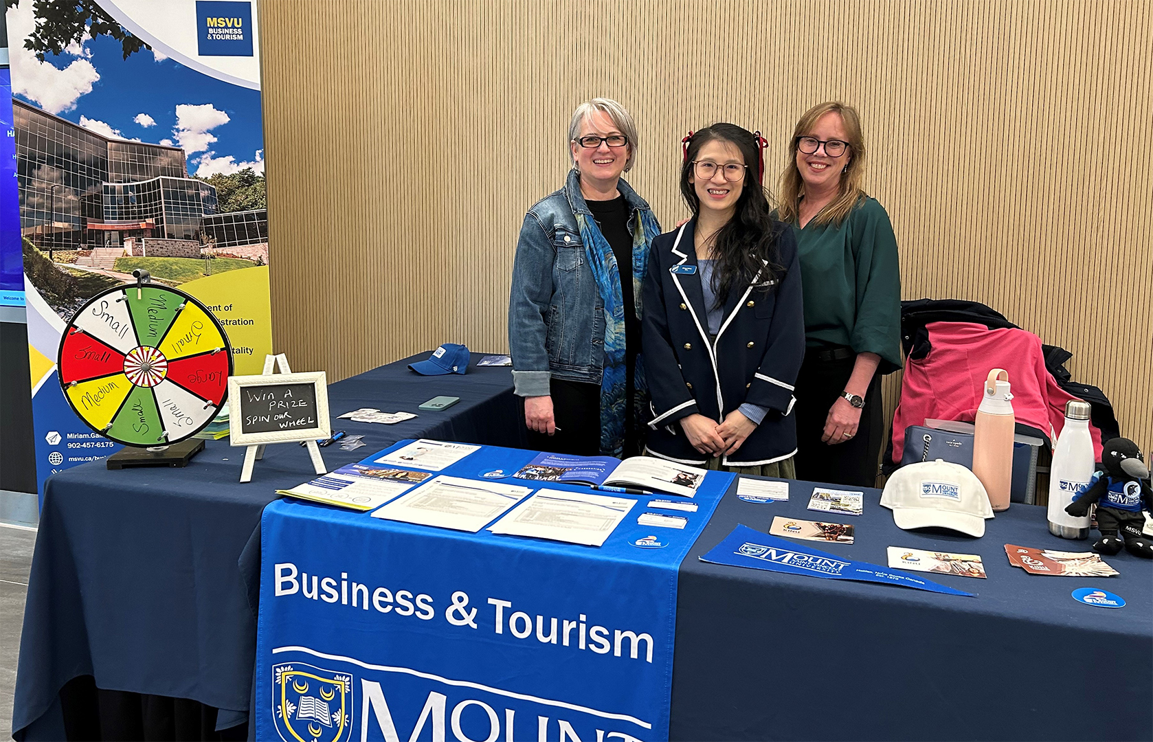 Two full time Tourism and Hospitality Management faculty and one student standing behind a table laid out with Mount gear at the hospitality fair in 2024.