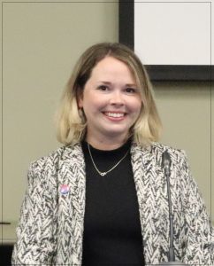 Catherine Chase, at the fall career week management panel session. Smiling, she has shoulder length blond hair, big smile, black shirt with grey and white patterned blazer on and a necklace. 