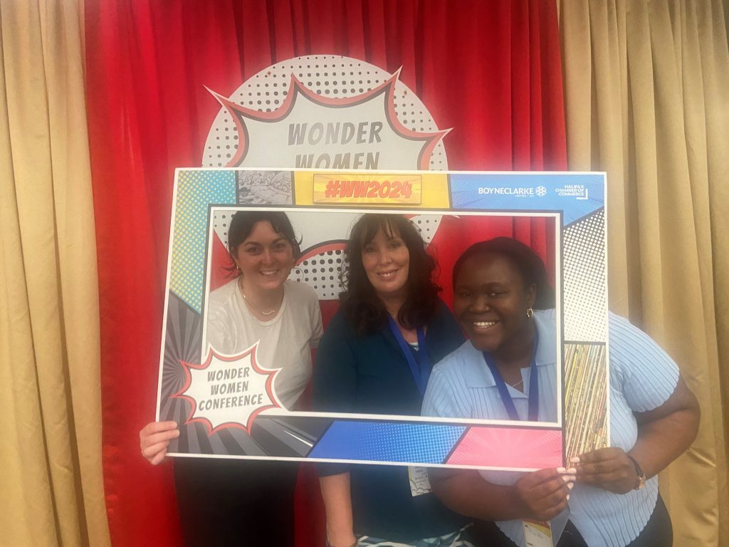 Wonder Women conference is a yearly event of the Halifax Chamber of Commerce. This year our Wonder Women students Precious and Megan and faculty Maria Matthews. These three are standing in front of a Wonder woman sign situated on a red and gold curtain. The three of them are holding a picture frame. 