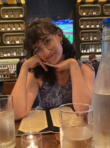 Lily is sitting at a restaurant table. She is wearing a blue top and has curly black hair and glasses.