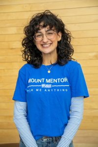 Lily is wearing a royal blue Mount Mentors Ask Me Anything t-shirt over a blue and white striped long-sleeve shirt, and glasses.