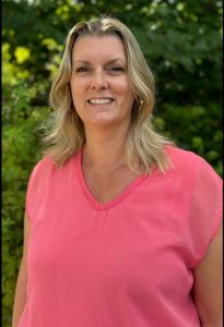 Nicolle is standing in front of a green bush. She is wearing a pink shirt, and has yellow hair and blue eyes.