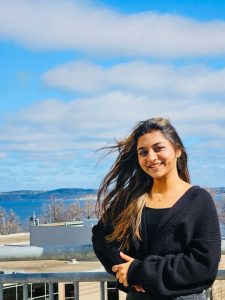 Simranpreet is leaning against a balcony railing wearing a black top and the blue sky is behind her.