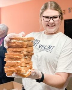 A student with glasses and a MSVU t-shirt, holding a stack of sandwiches